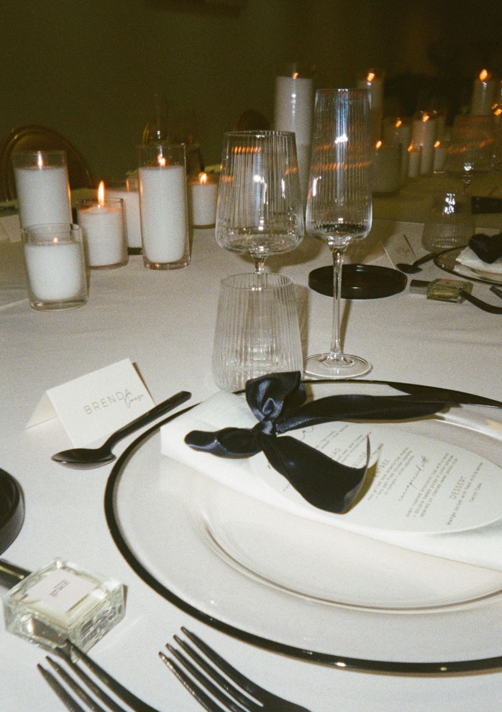 Place settings at an elegant dinner party feature white plates, black chargers, wine glasses and glowing candlelight.