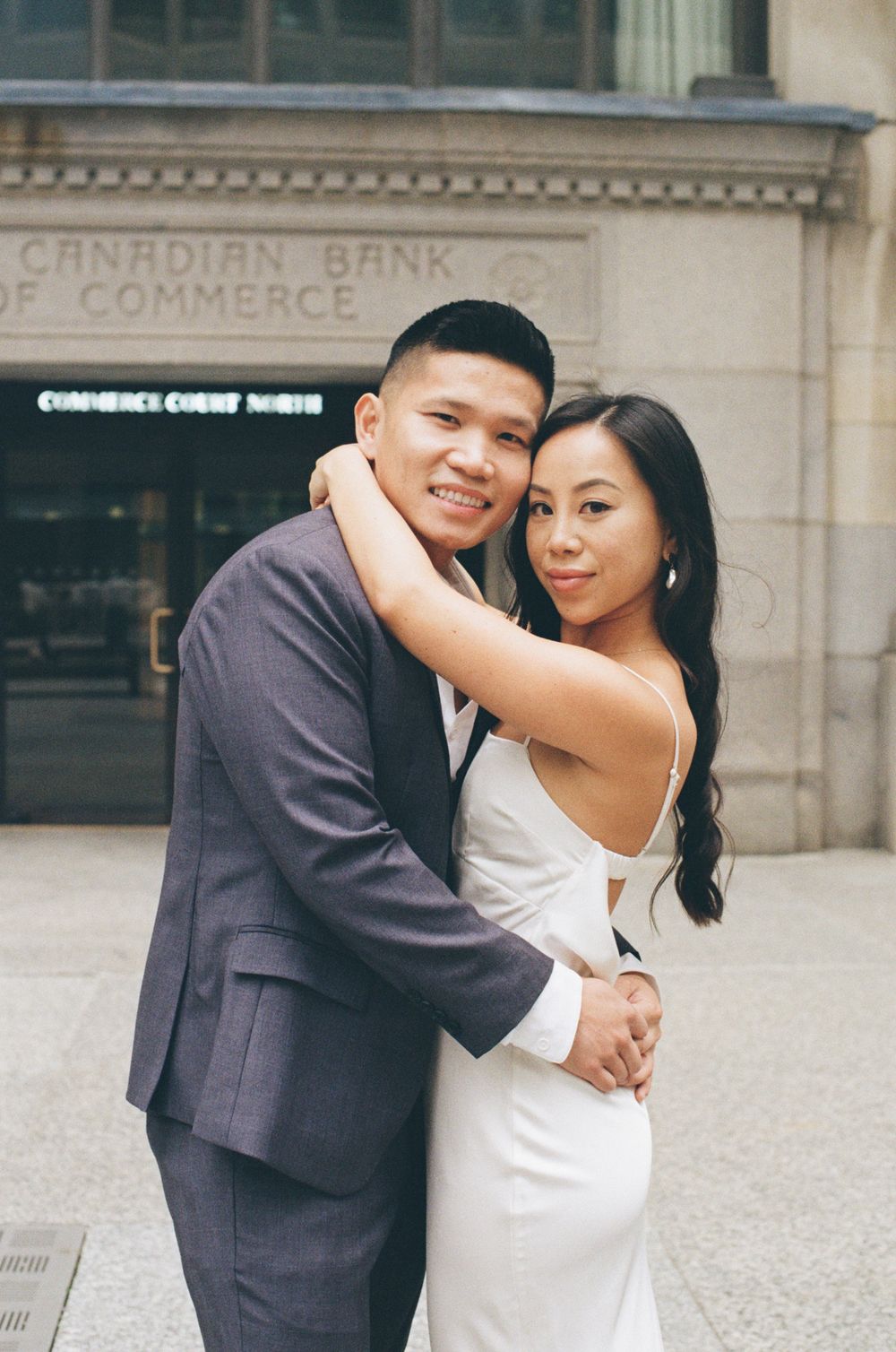 A romantic wedding couple embraces in elegant formal attire outside a classical stone building.