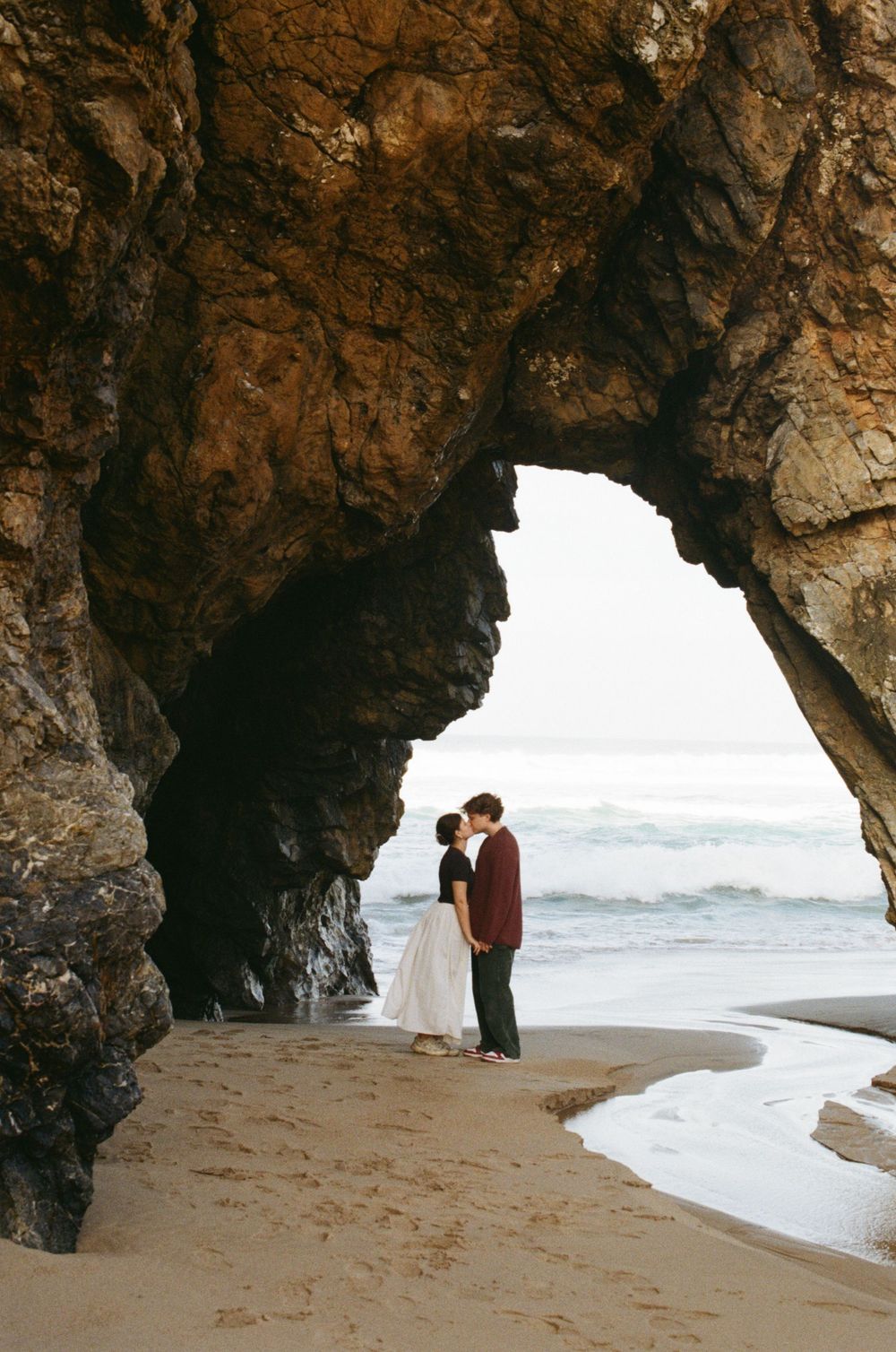 A couple shares a romantic moment under a natural stone arch at a beach during sunset with crashing waves in the background.