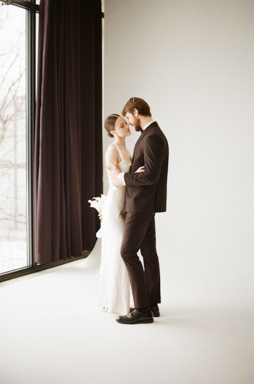 A couple shares an intimate dance pose near a bright window while wearing formal wedding attire.