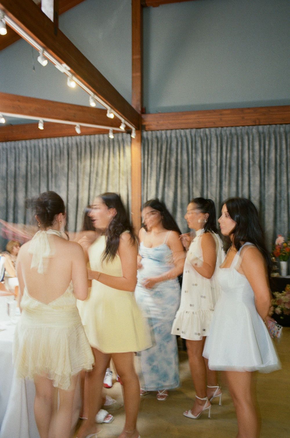 A group dances and celebrates in a rustic indoor venue with string lights and grey curtains in the background.