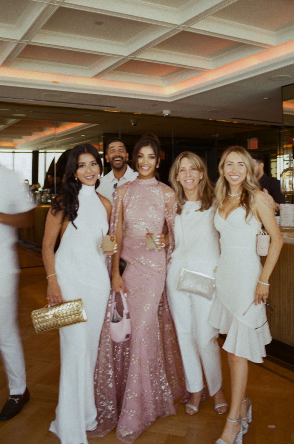 Group of friends wearing elegant white and pink sparkly dresses pose together at an indoor celebration.