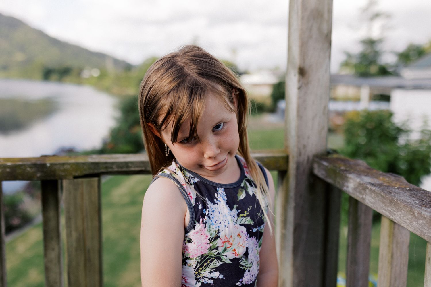 A child wearing a floral navy dress poses on a wooden deck railing with mountains visible in the background.