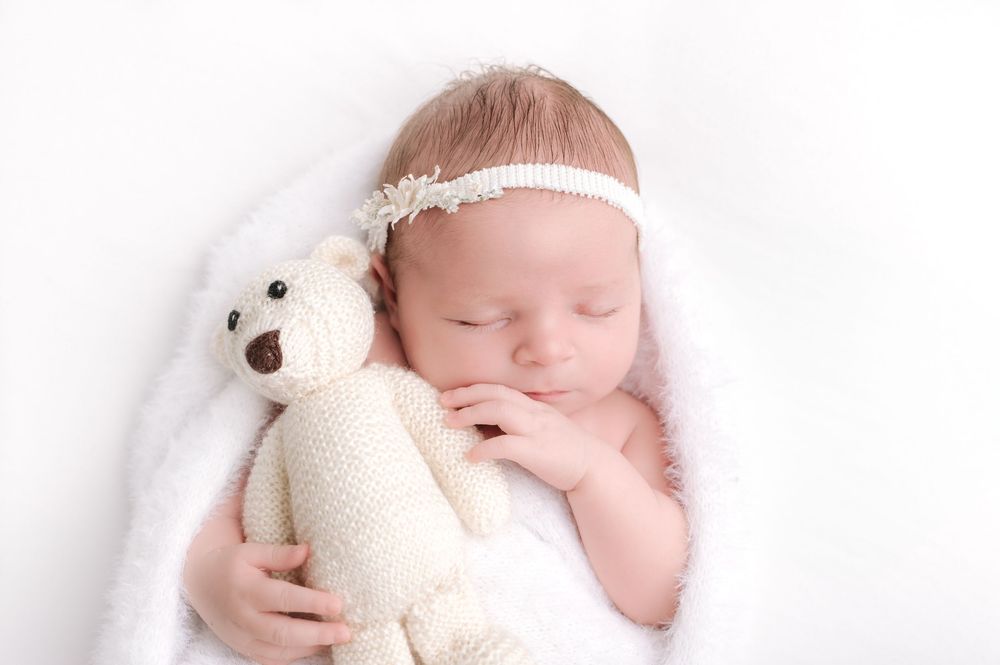 Newborn baby sleeping peacefully wrapped in white blanket, holding cream knit teddy bear, wearing floral headband.