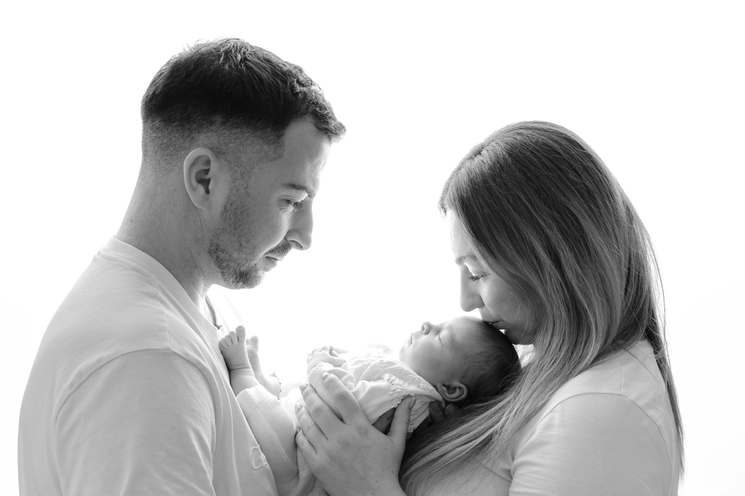 Black and white photo of parents lovingly gazing at their newborn baby in a bright studio setting.
