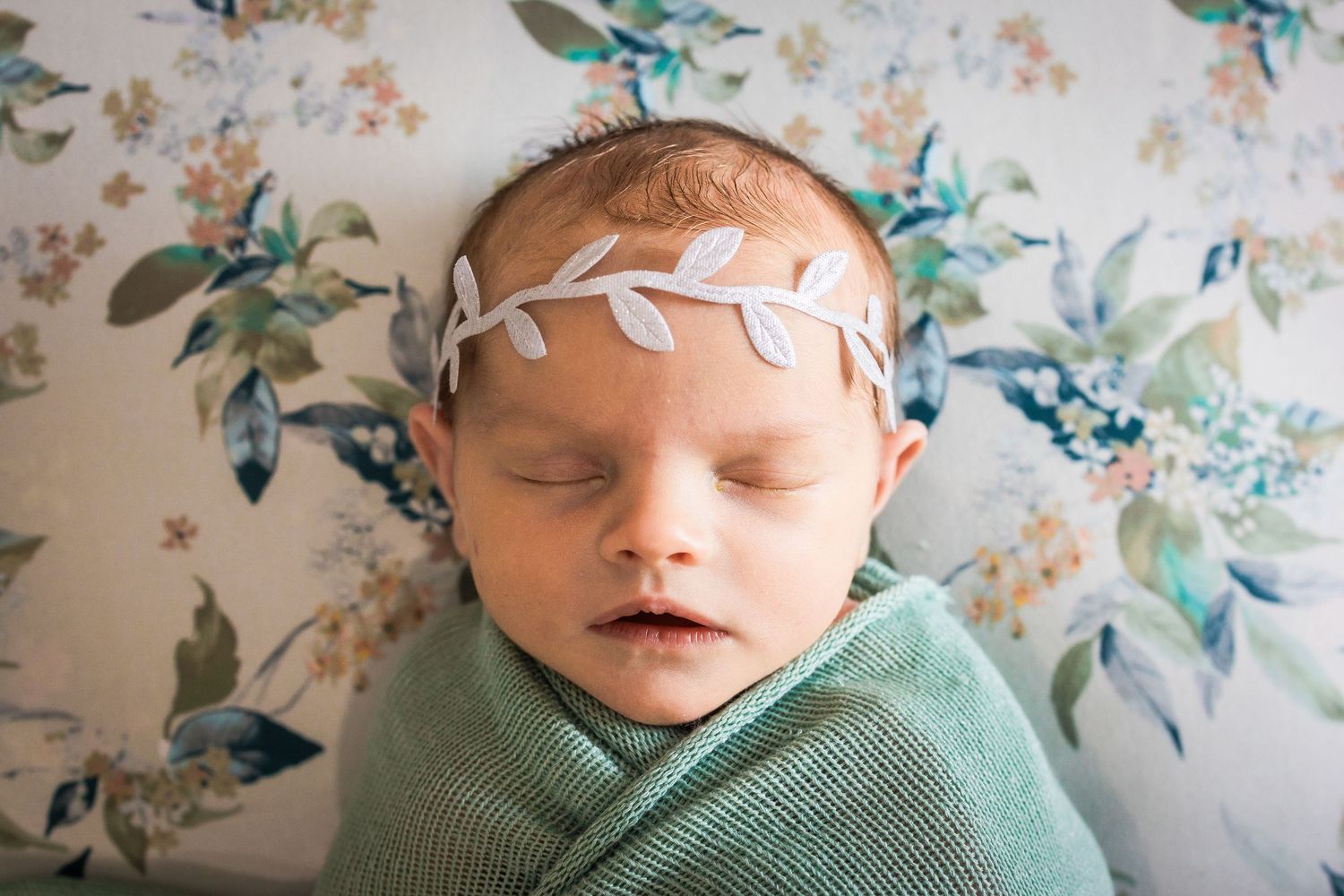 Newborn baby wrapped in mint green swaddle against floral background.