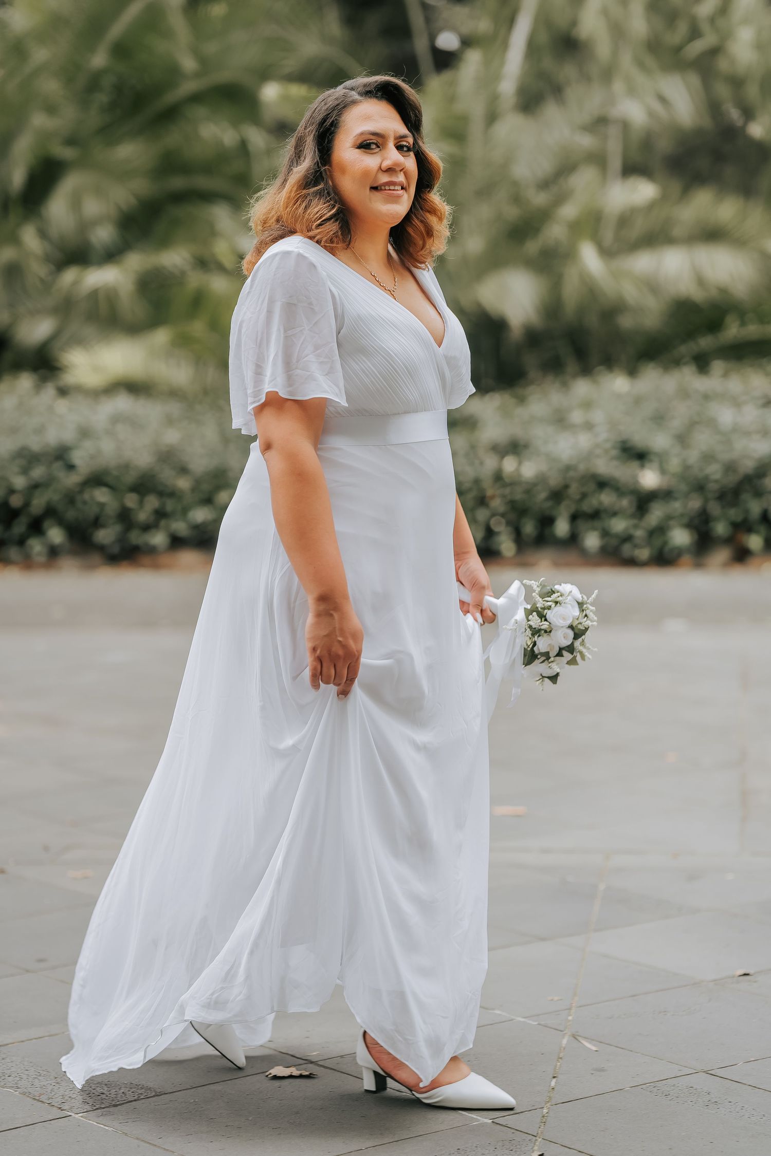 A bride in a flowing white dress holds a small bouquet while posing outdoors in a garden setting.