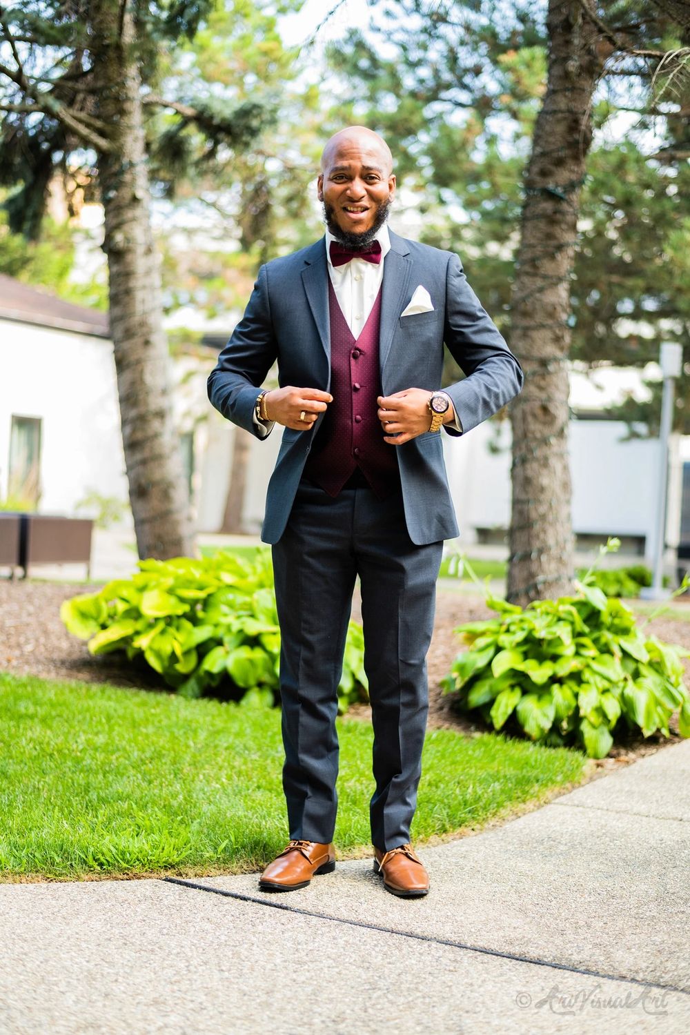 Dapper gentleman in navy suit and burgundy bow tie poses outdoors on garden path.