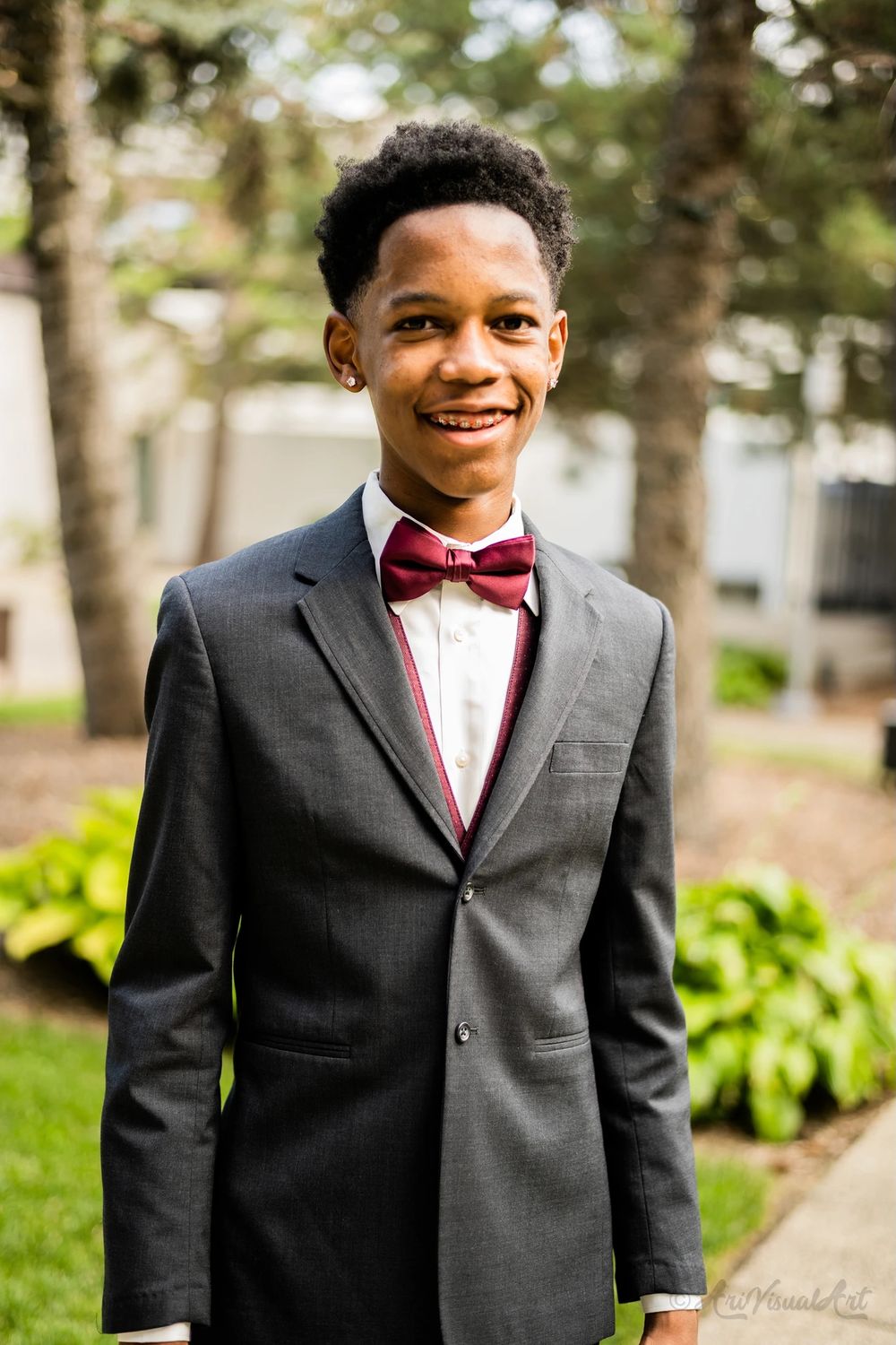 Young man in classic charcoal suit and burgundy bow tie smiles in outdoor setting.