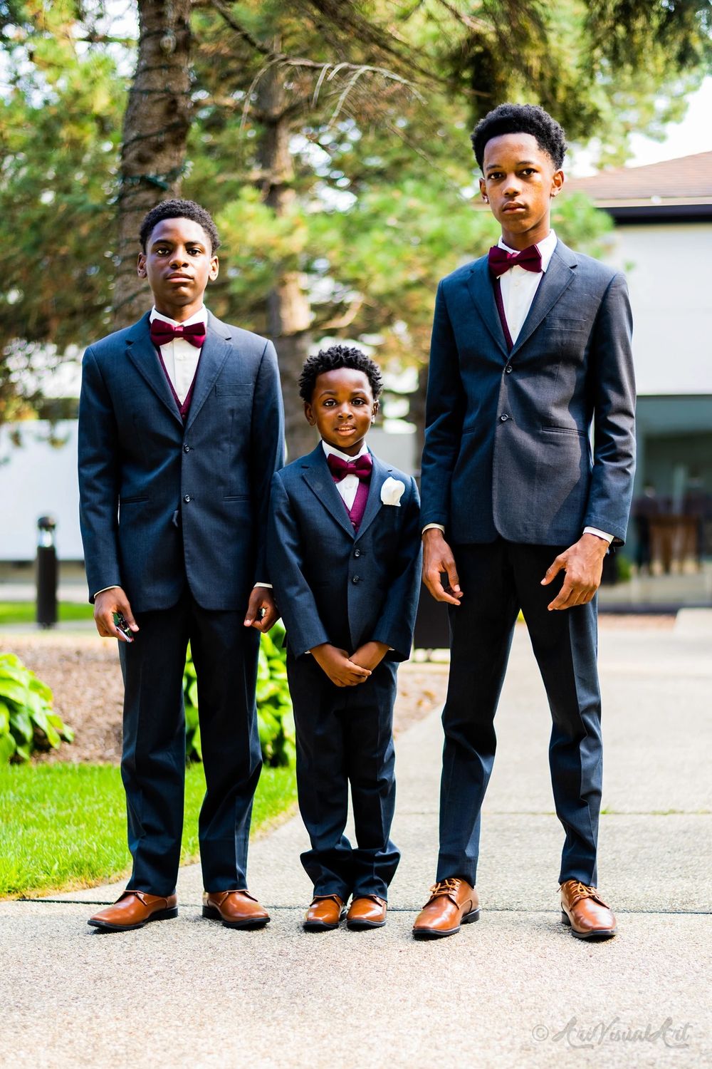Three dapper groomsmen in matching navy suits and burgundy bowties stand together on a garden path.