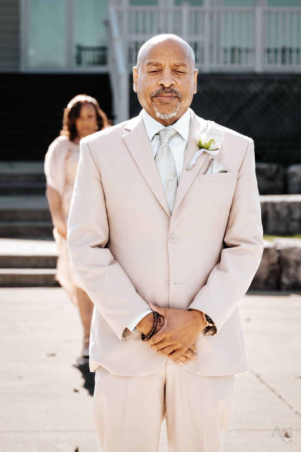 A wedding guest in a beige suit with boutonniere poses confidently outdoors.