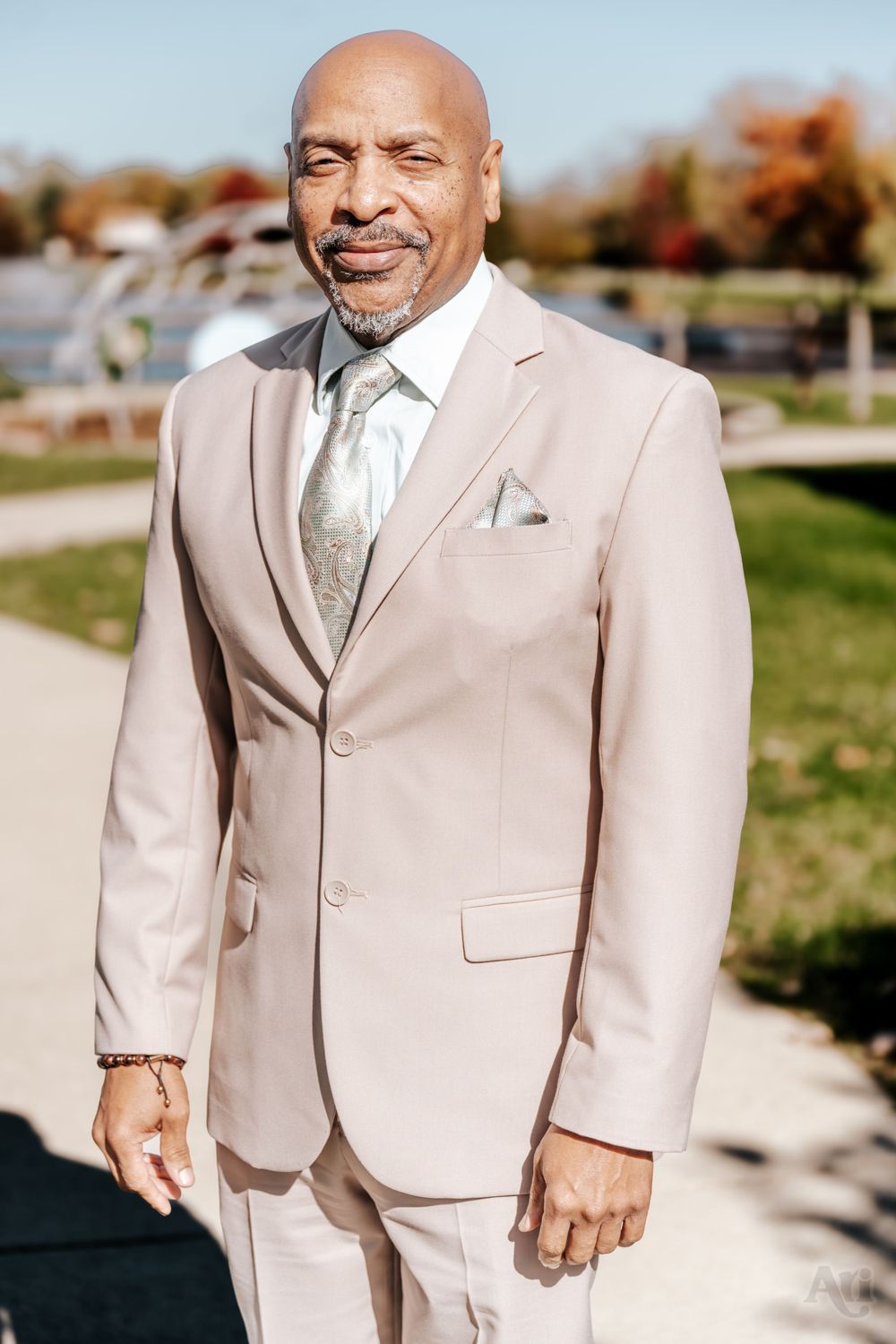 A wedding guest in a light beige suit and patterned tie smiles warmly outdoors.