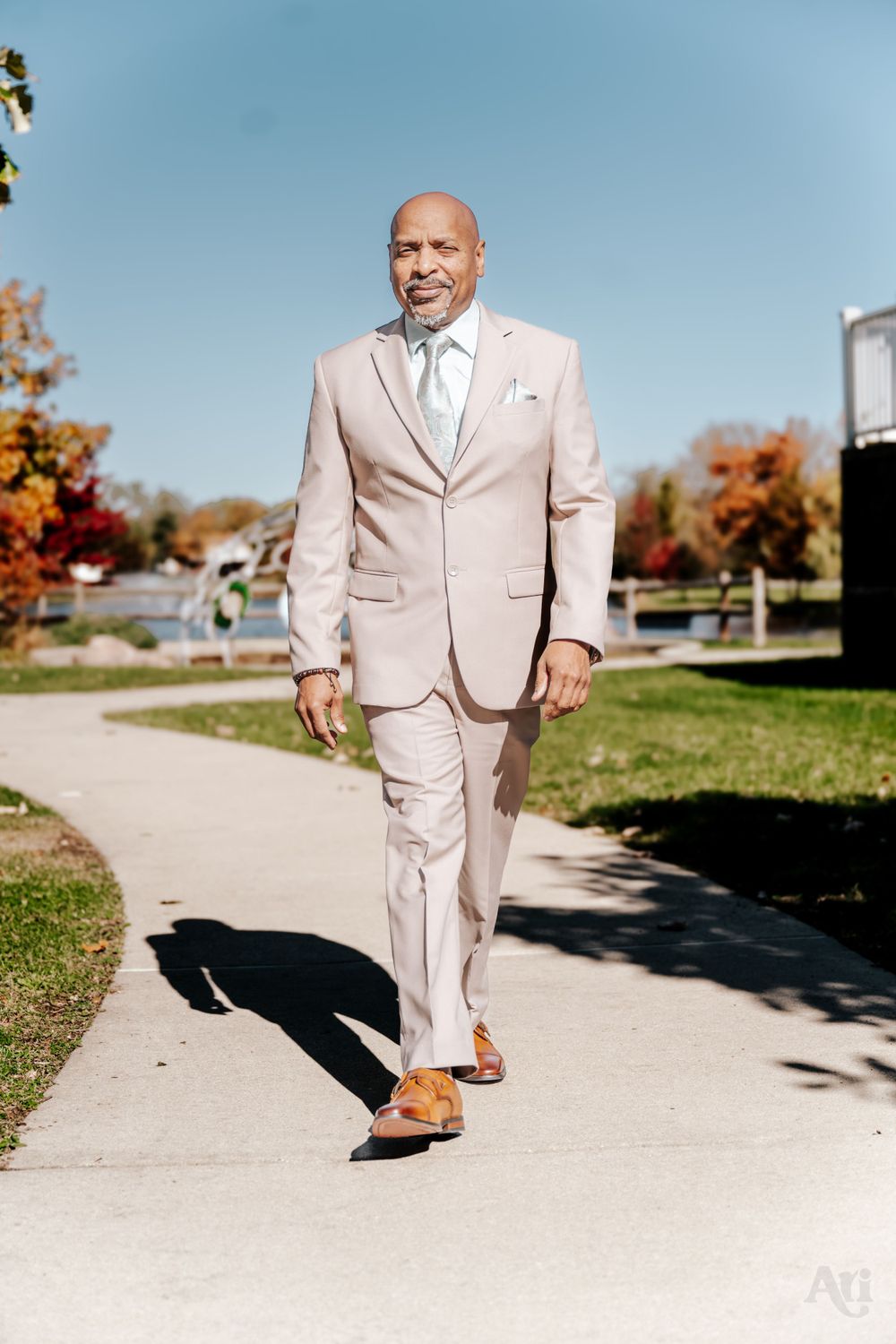 A wedding guest in a light beige suit walks confidently along an outdoor path.