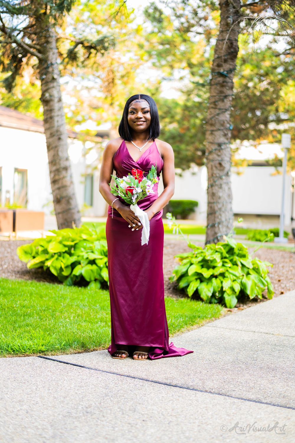 A bridesmaid in a burgundy gown holds a floral bouquet while standing on a garden pathway.