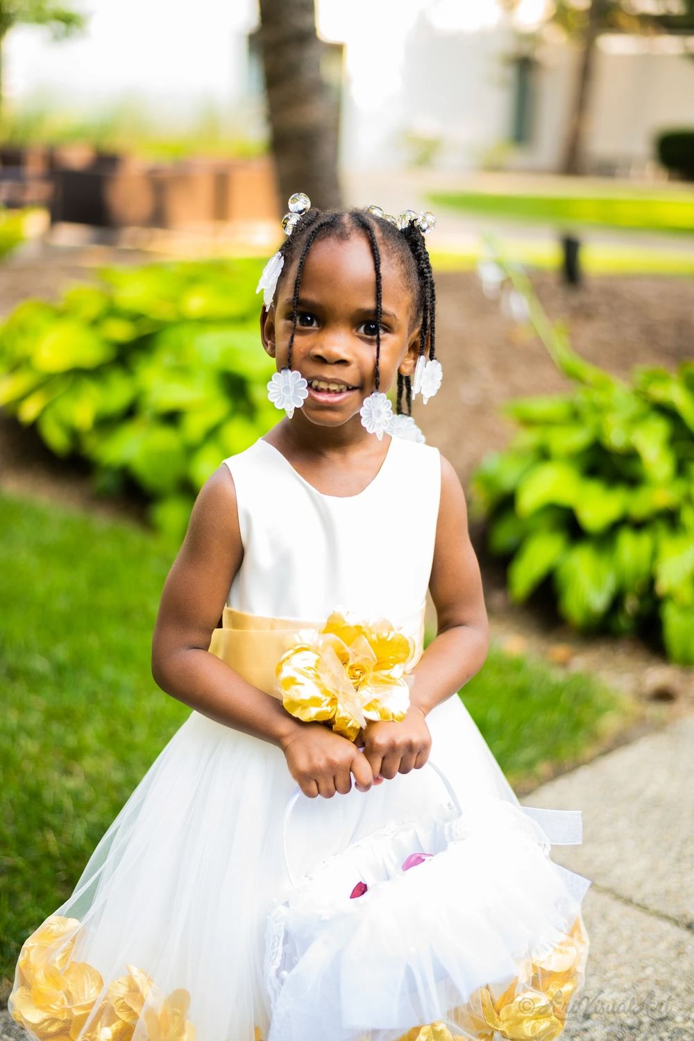 A flower girl in a white and gold dress smiles brightly while holding flowers.