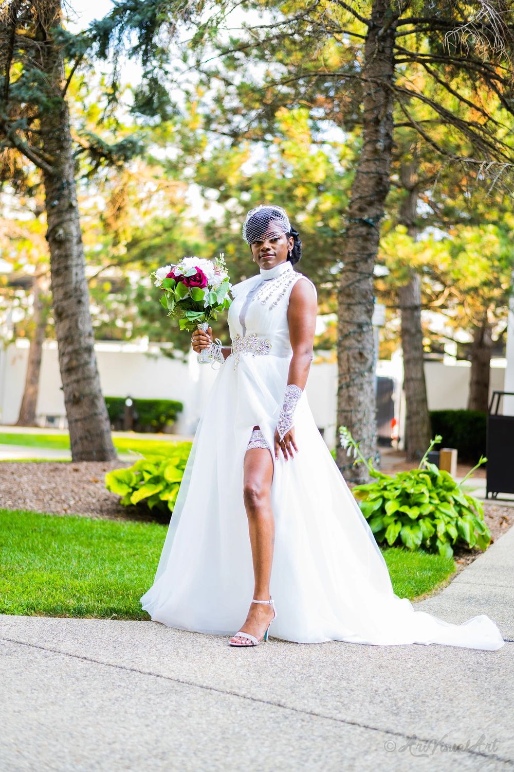 A bride in an elegant white gown with high slit poses with bouquet on a garden path.