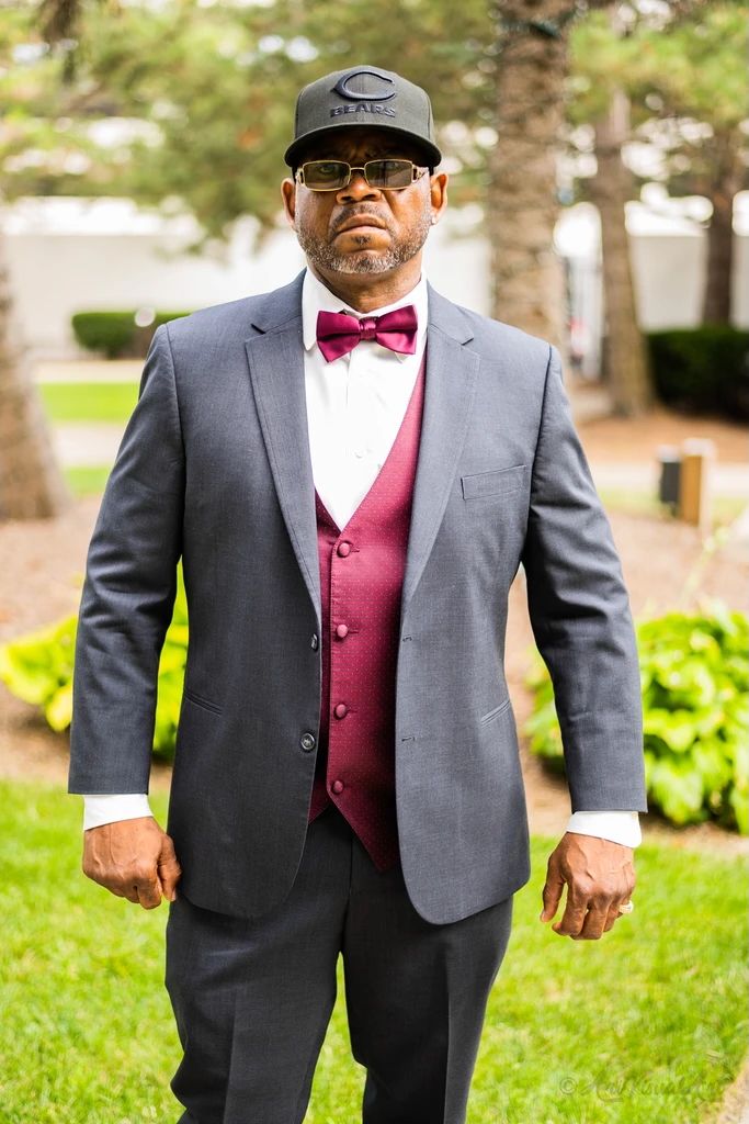 A wedding guest in a gray suit with burgundy vest and bowtie poses outdoors.