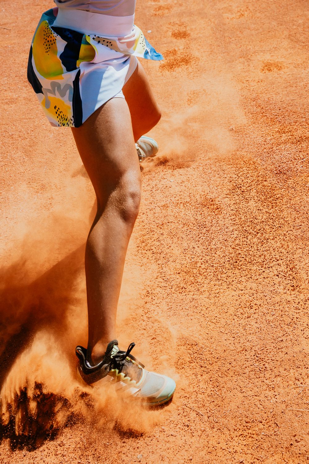 Tennis player's legs kicking up red clay dust on court during match action with white shoes and colorful shorts.