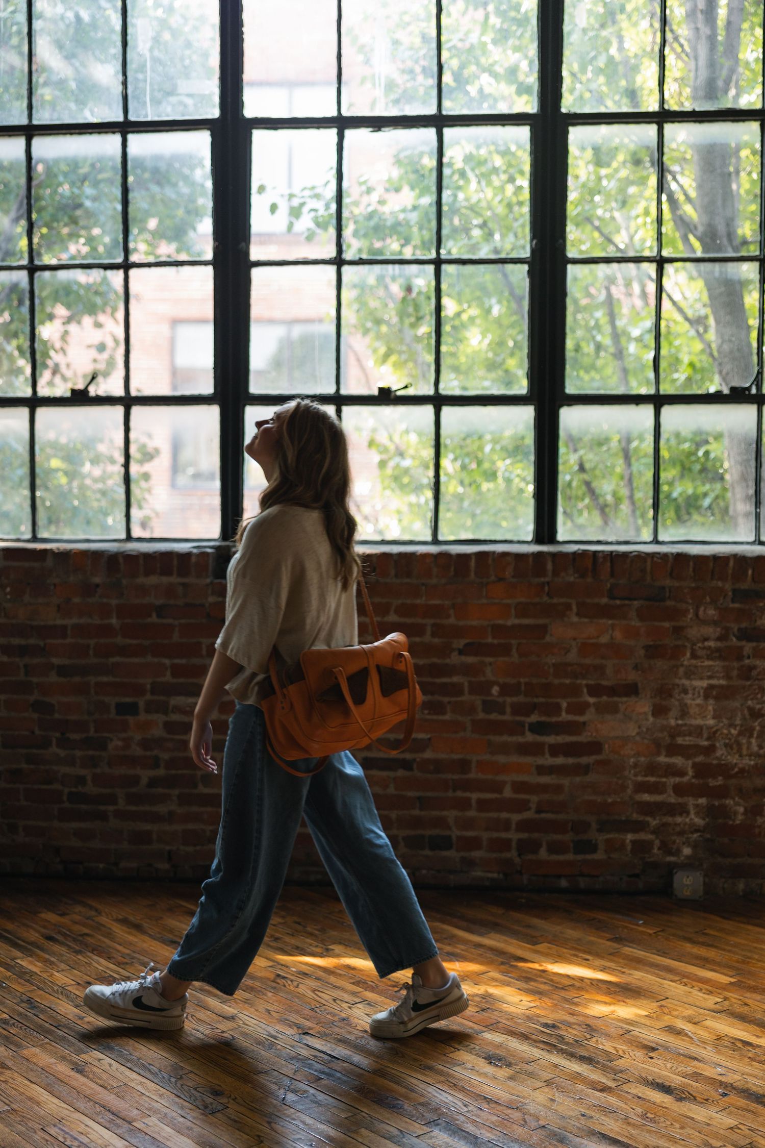 A photographer walks on a wooden floor in a sunlit studio with exposed brick walls and large windows.