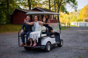 Brides waving from golf cart with guest at autumn wedding venue.