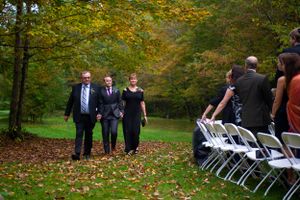 Bride escorted down aisle by family members at outdoor fall wedding ceremony.
