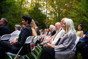 Wedding guests seated in white chairs enjoying outdoor autumn ceremony in woods.