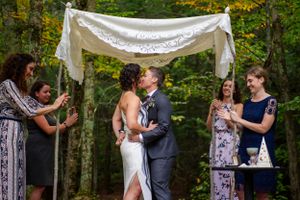 Couple shares first kiss under white lace chuppah at outdoor woodland wedding ceremony.