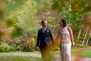 Newlywed couple walks hand in hand through colorful autumn foliage at outdoor wedding venue.
