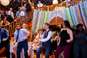 Wedding guests dance energetically inside rustic barn venue decorated with colorful ribbon backdrop.