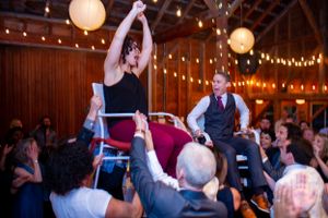 Newlywed couple lifted in chair during lively hora dance at rustic barn wedding reception celebration.