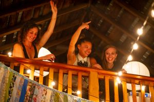 Three young women laughing and waving from a wooden balcony decorated with string lights.