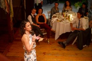 Woman in floral dress speaking to seated wedding guests in warmly lit rustic venue.