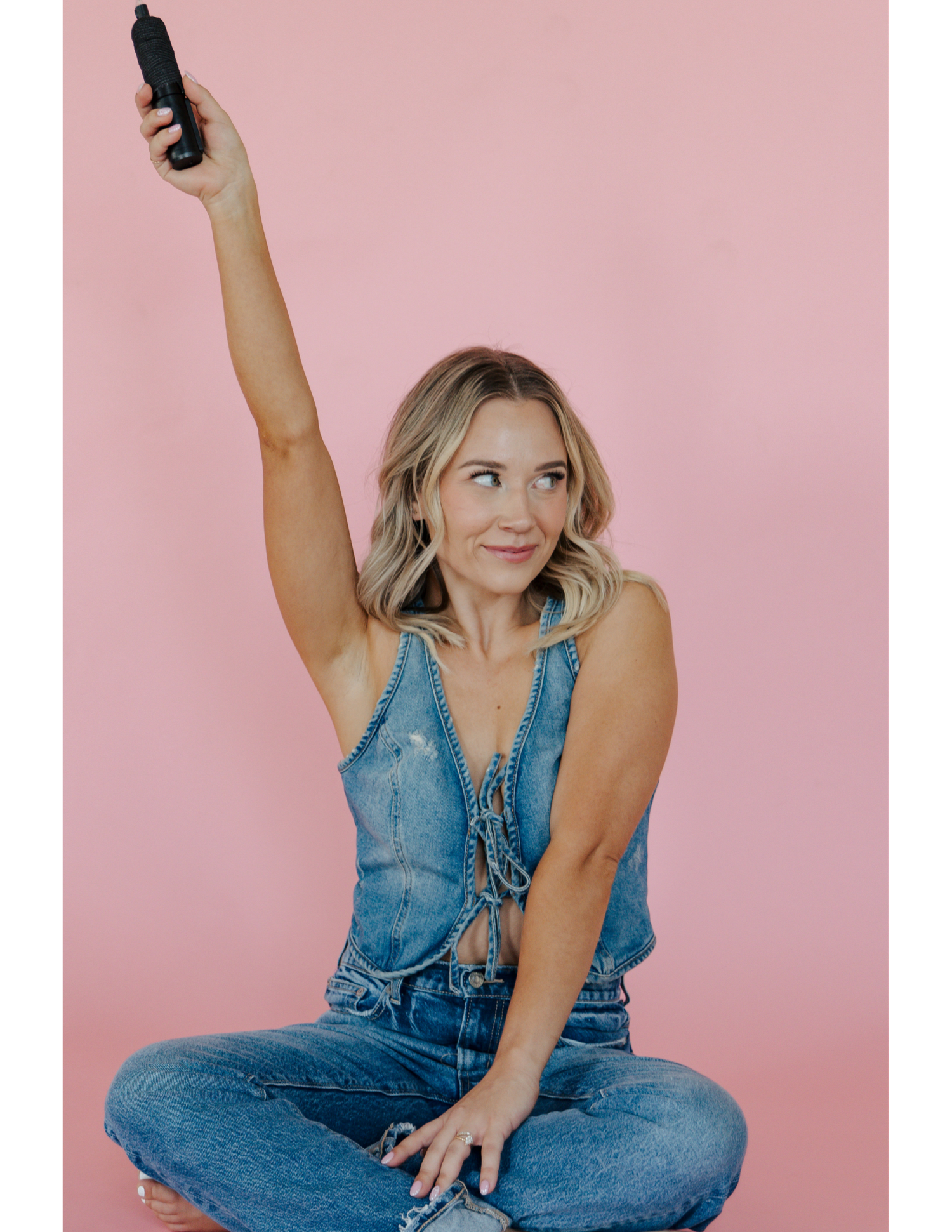 Woman in denim outfit sits cross-legged and raises a phone while smiling against pink background.