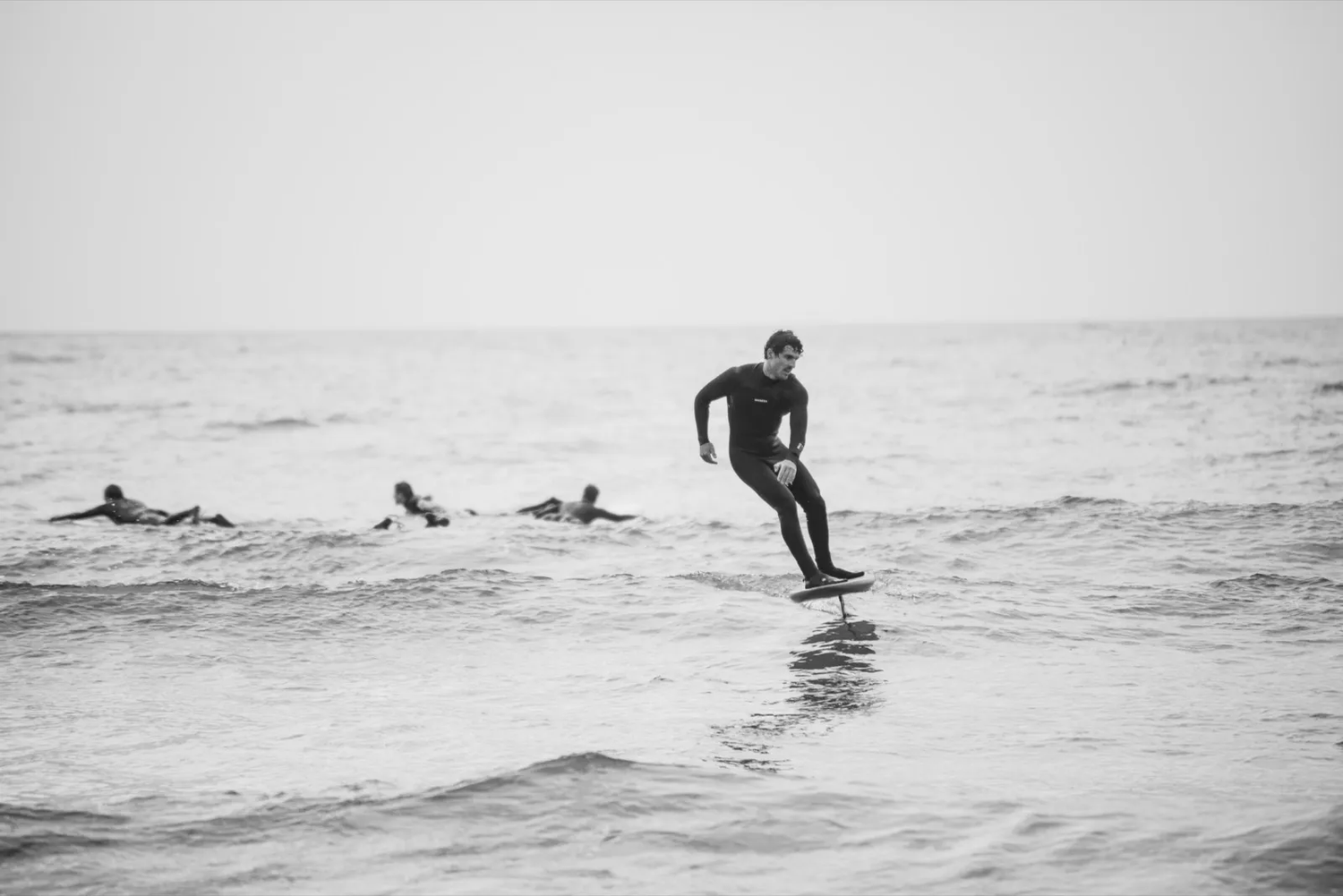 Un surfeur glisse sur une vague tandis que plusieurs autres attendent à l’arrière-plan, dans une ambiance brumeuse et monochrome.