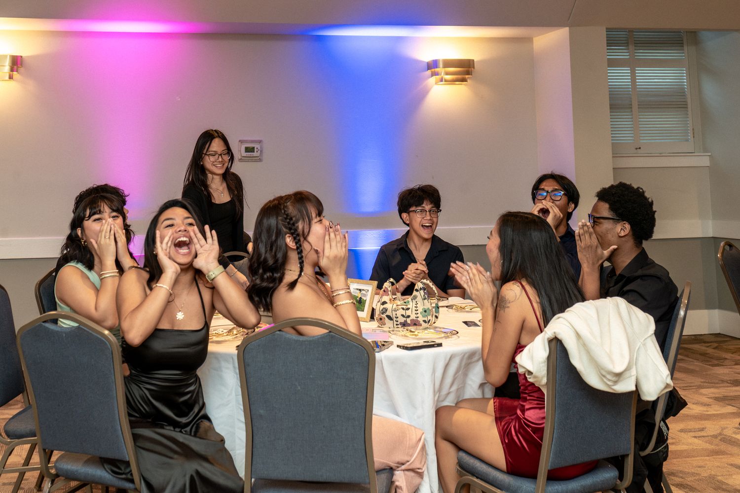 A group of people sitting at a long dining table enjoying a meal together under colorful mood lighting.