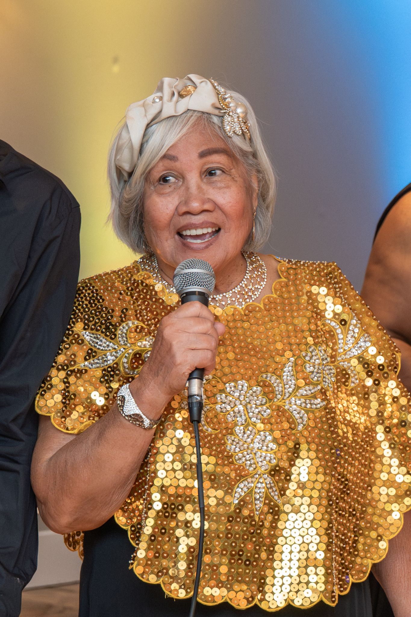 A performer in a dazzling gold sequined outfit and white headpiece sings into a microphone during a show.