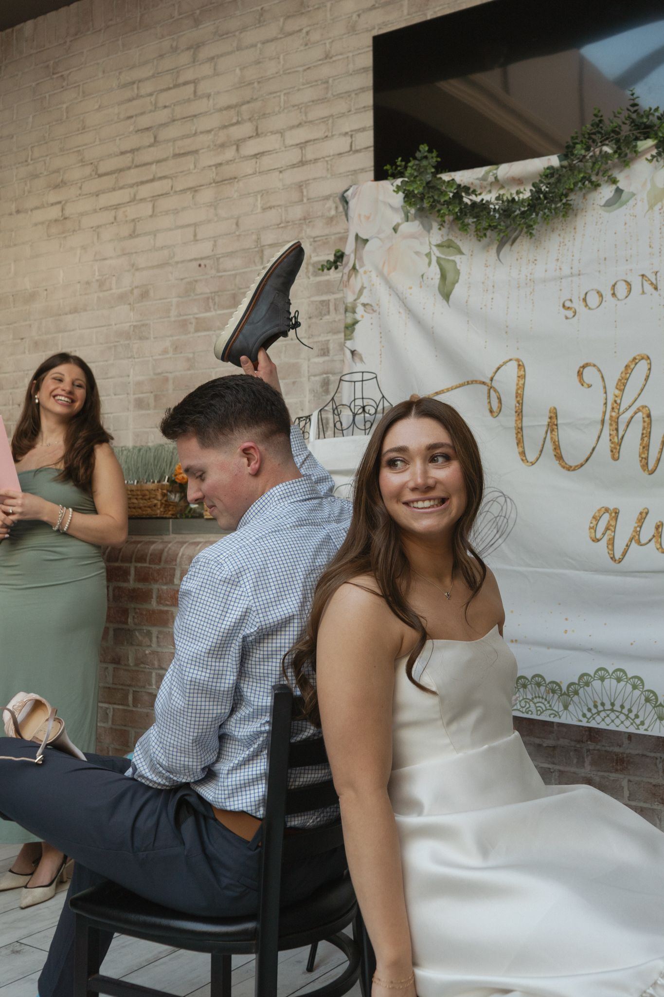 Couple celebrating at a wedding shower with white brick wall backdrop and 'Soon' banner with greenery decorations.