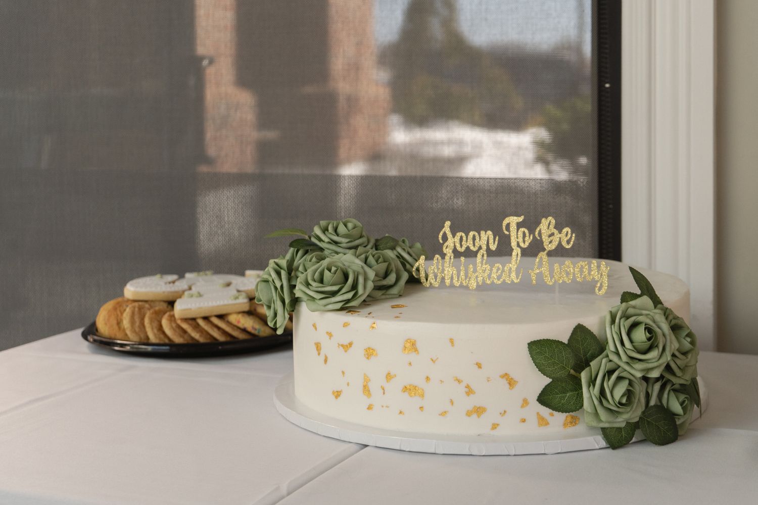 A white wedding cake decorated with green floral accents sits on display at an elegant reception venue.