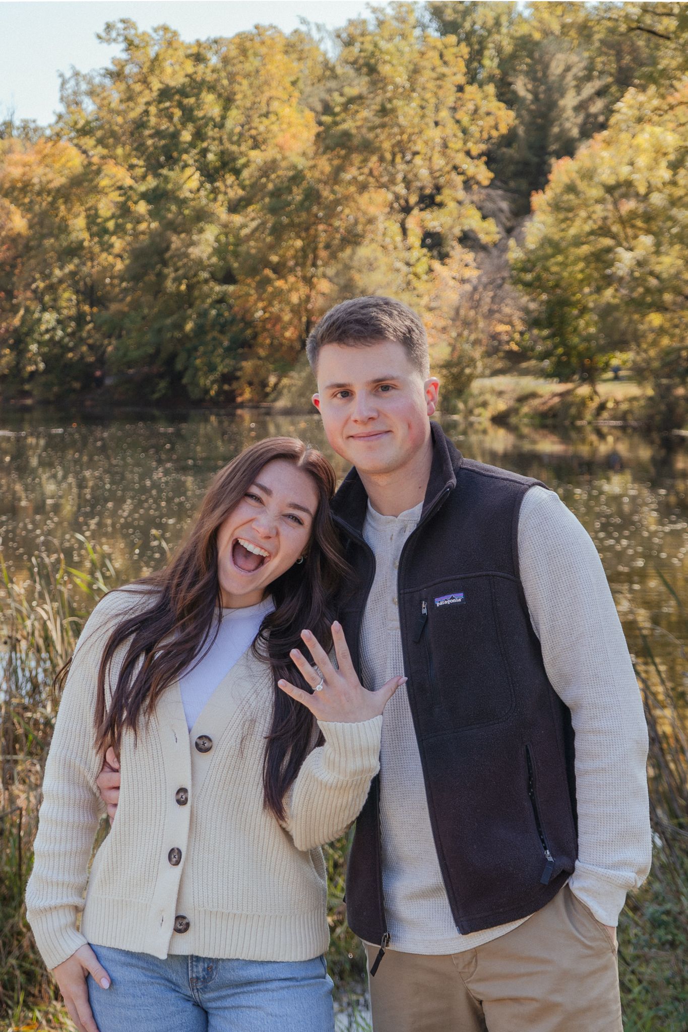 A couple in fall attire poses outdoors showing off an engagement ring amid autumn foliage.