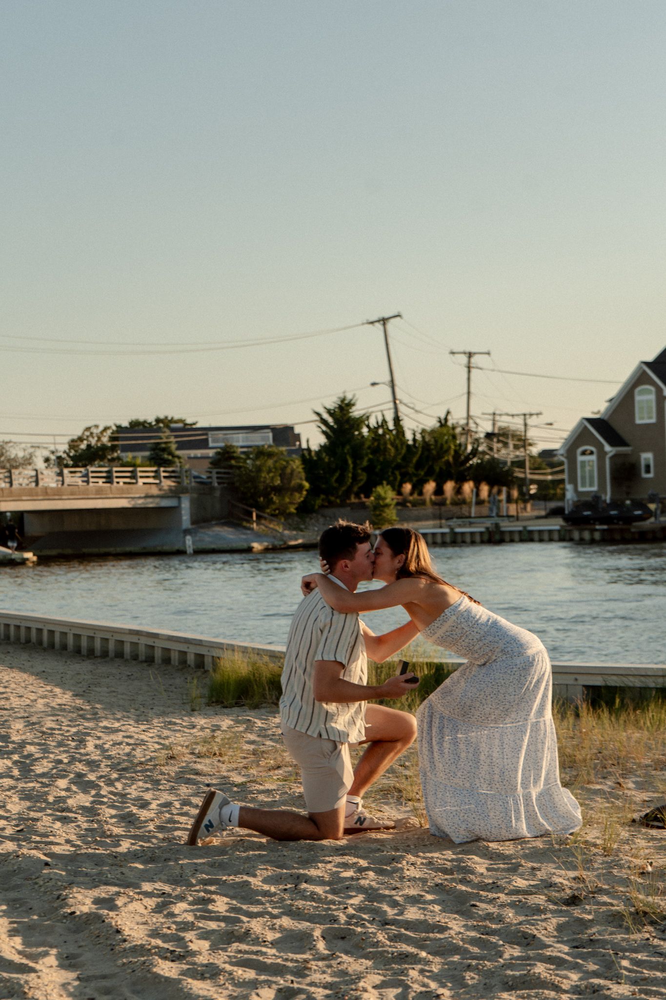 Couple sits together on sandy beach at sunset with coastal houses and water in background.