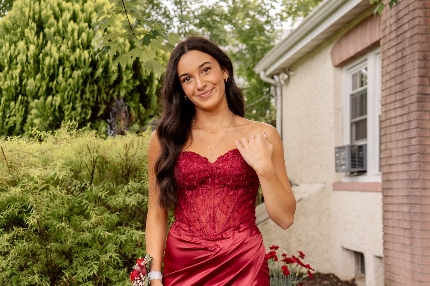Person in a burgundy formal dress posing outdoors near a residential building.