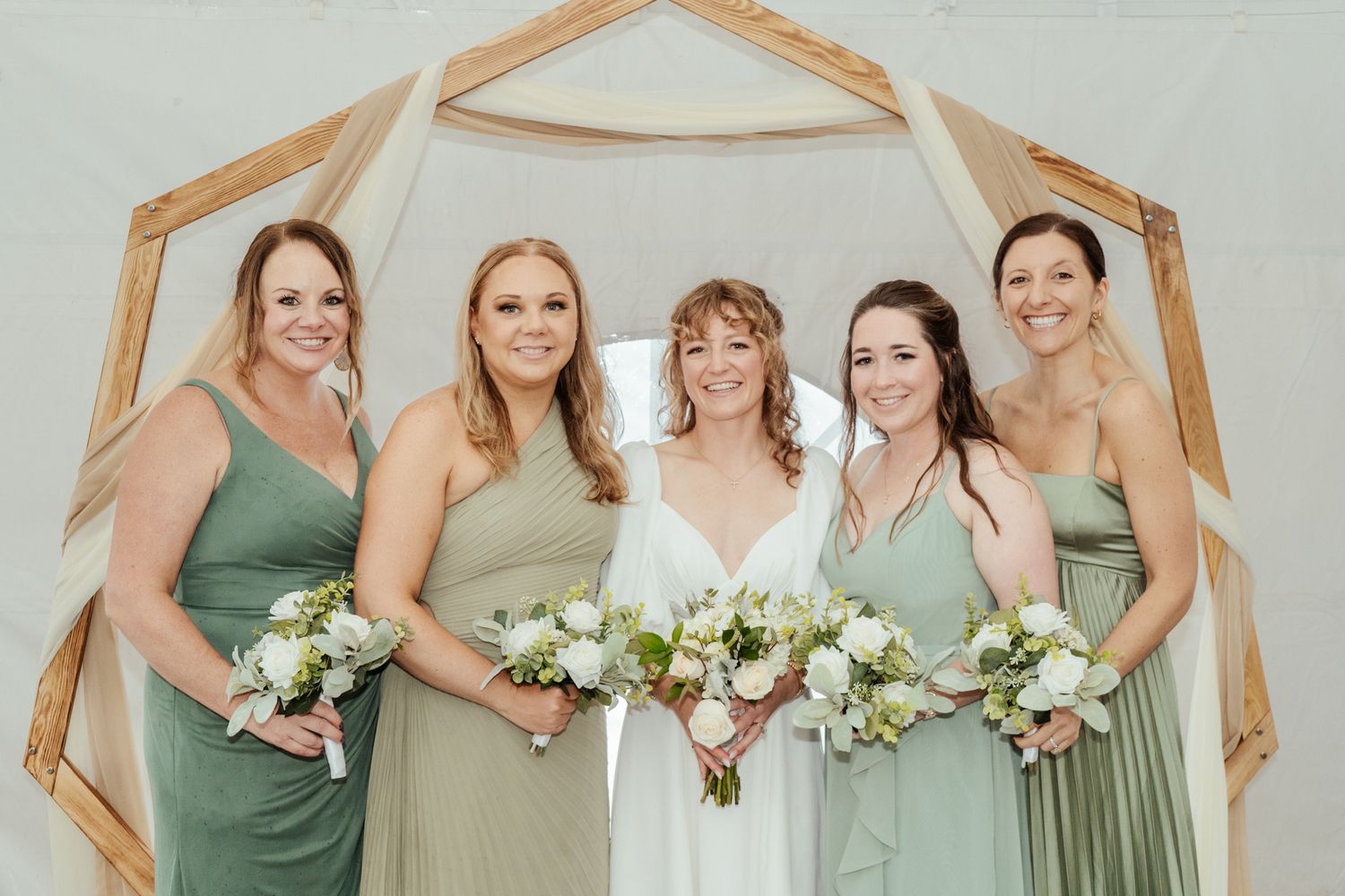 Bridesmaids in sage green dresses pose together holding white floral bouquets.
