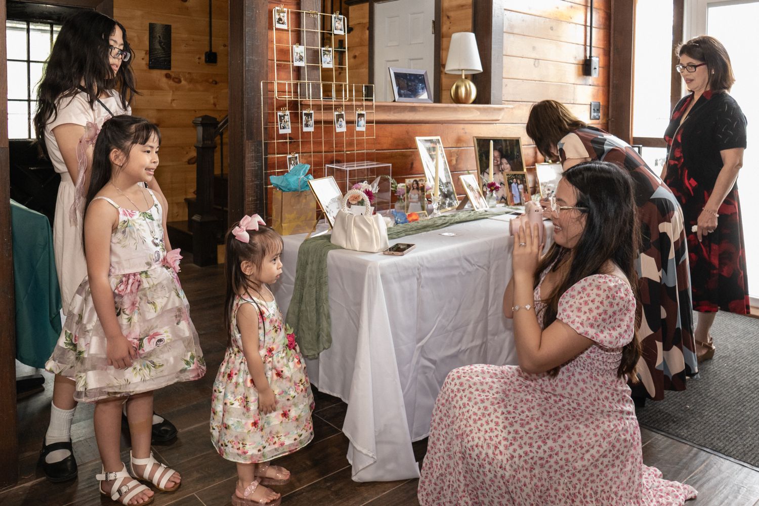 Wedding guests in floral dresses gather at an indoor venue with wooden beams and natural lighting.