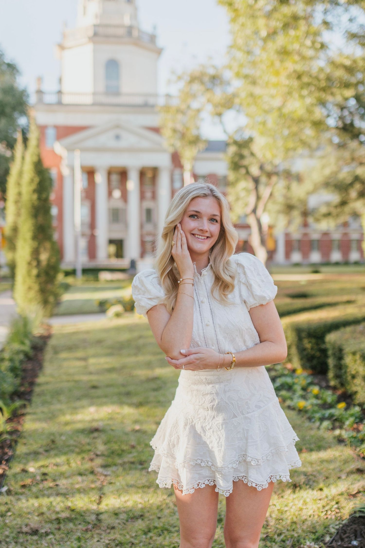 Catherine Kramer, Dallas portrait photographer, smiles in a white lace outfit and poses in front of a classic brick building.