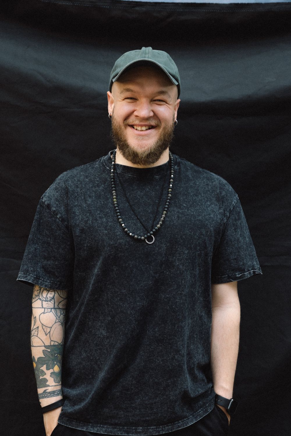 Person in dark t-shirt and baseball cap with arm tattoos smiling against black backdrop.