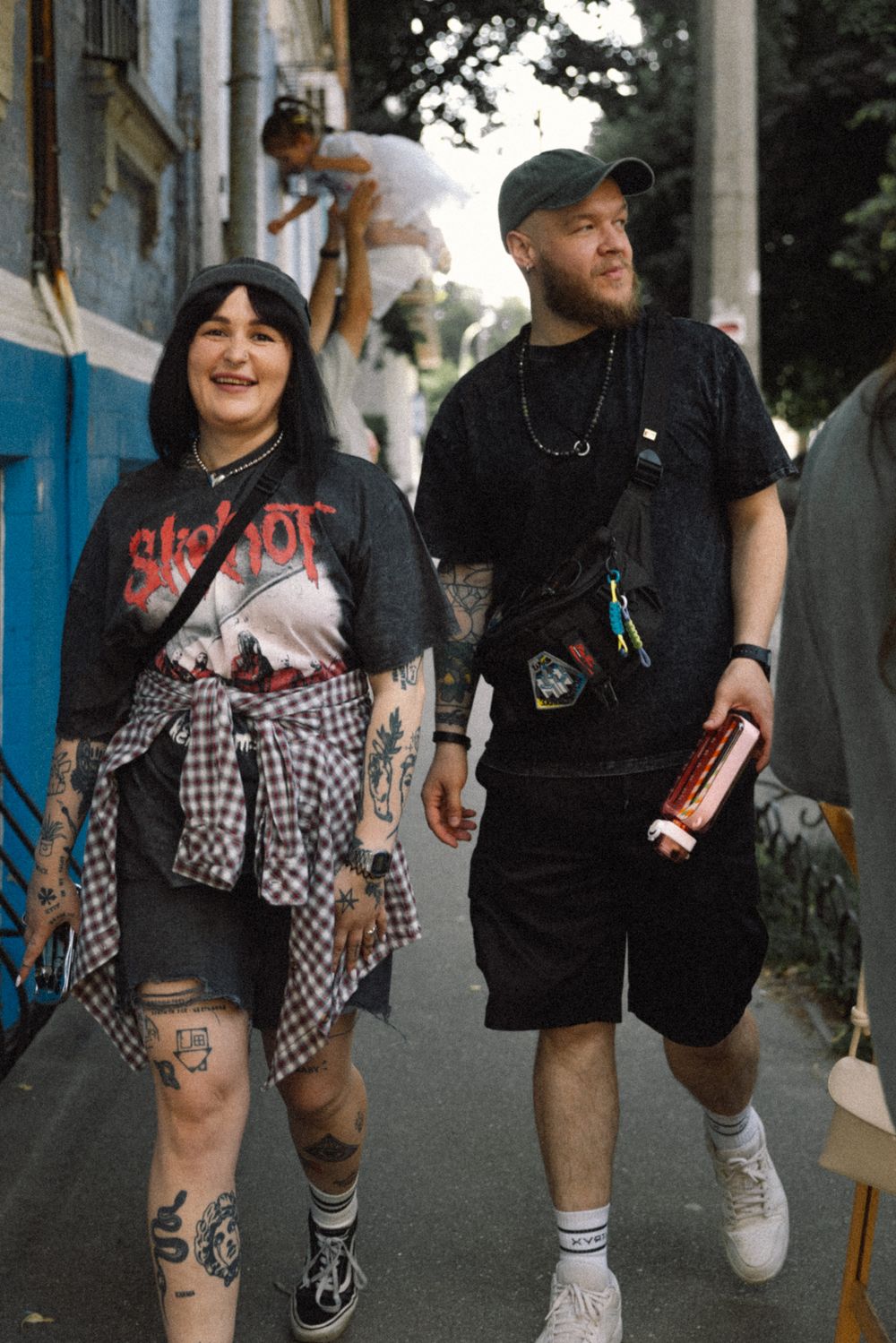Two smiling figures in black clothing and checkered patterns walk down a sidewalk on a sunny day.