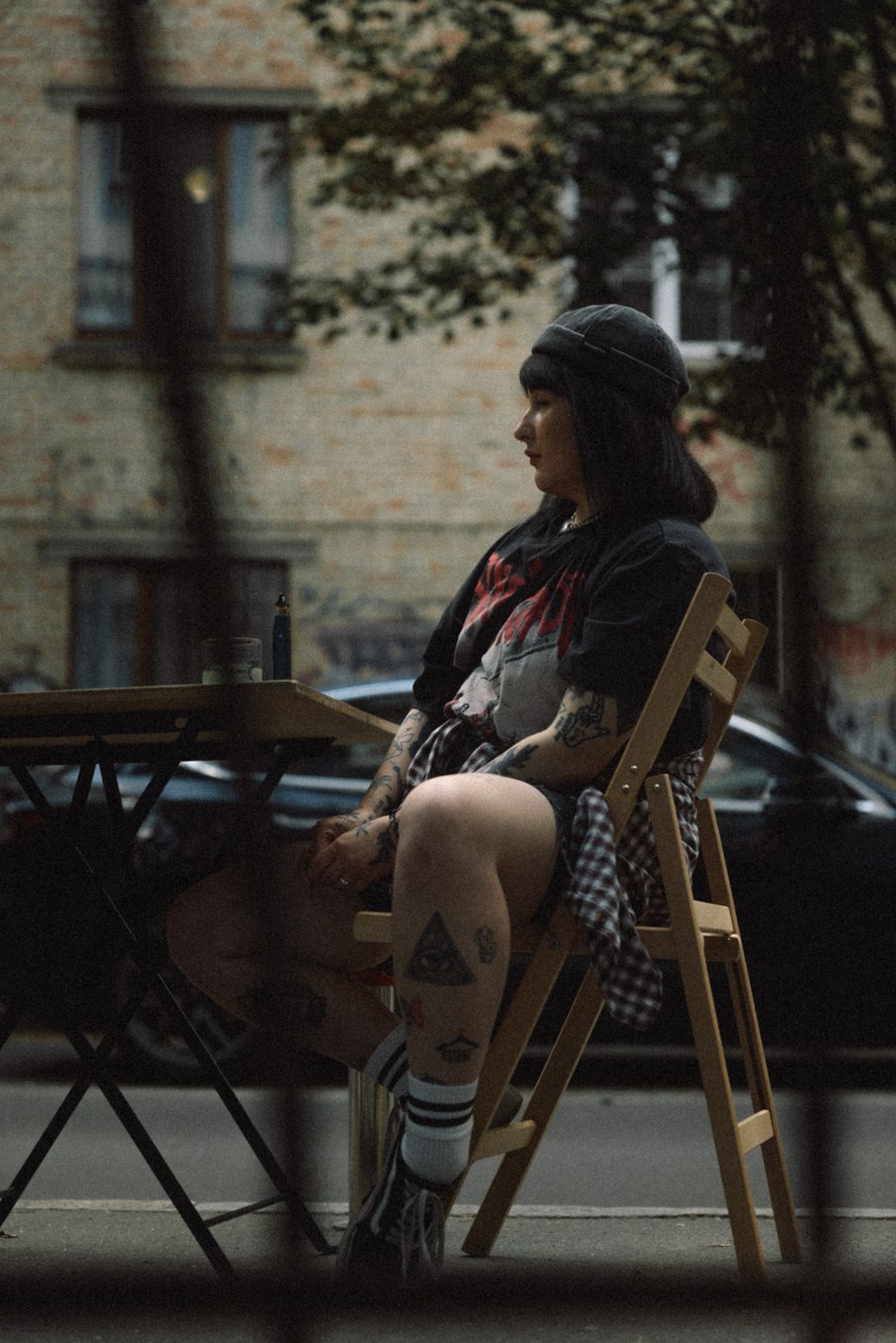 A person wearing a beanie and knee highs sits on a wooden folding chair on a city street during an overcast day.