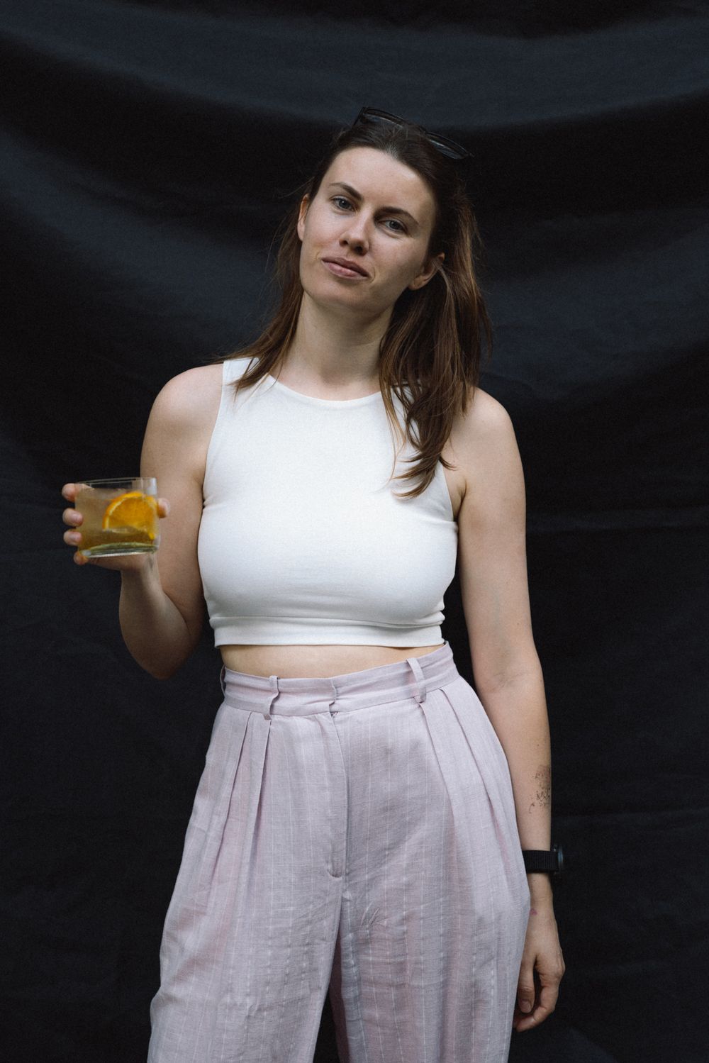 Figure in white crop top and pink high-waisted pants holds orange beverage against dark background.
