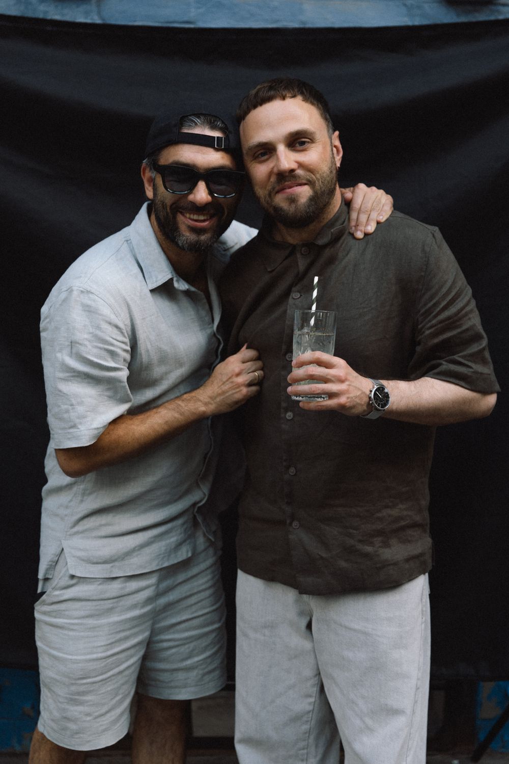 Two friends posing together at a casual outdoor gathering, one wearing sunglasses and light clothes, other in dark shirt.