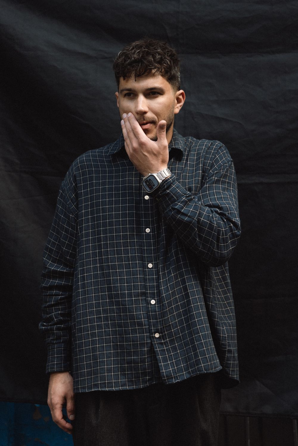A person wearing a dark plaid button-up shirt stands against a black background while touching their face thoughtfully.